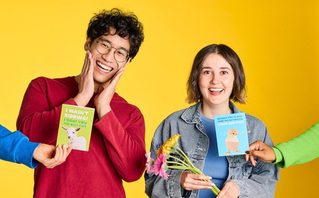 Two young adults smiling and laughing against a bright yellow background, each holding a colorful card. A person on the left in a red sweater holds a green book and touches their face, while a person on the right in a denim jacket holds a blue book and flowers. Photo: Michelle Jarni