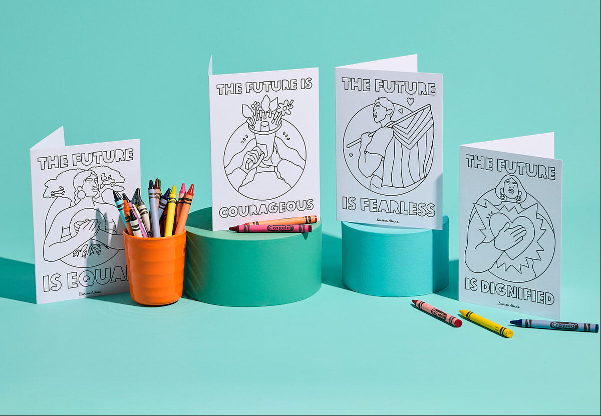 Four white greeting cards with black line-art illustrations and text reading 'THE FUTURE IS' followed by equality, courage, peace, and dignity, arranged on teal and mint display blocks with crayons in an orange cup and on the surface against a mint background. Photo: Michelle Jarni